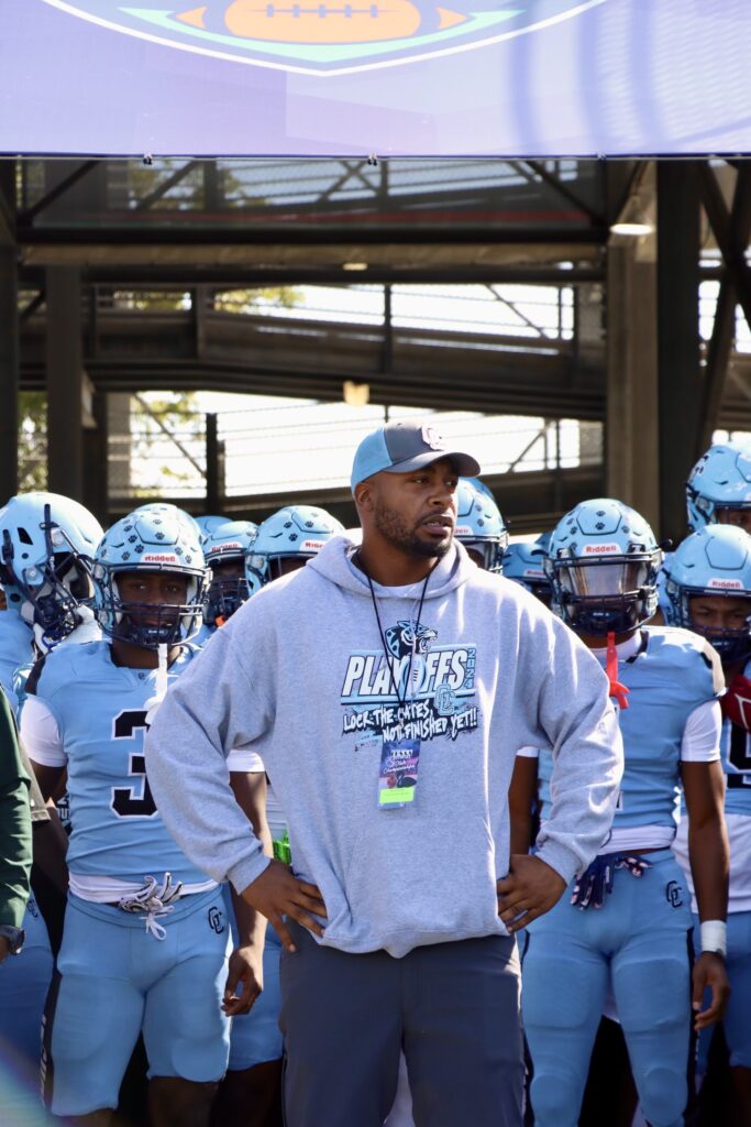 Embattled head coach Russell Ellington stands in front of his team, wearing a light blue and gray playoff sweatshirt, looking forward with determination as his players in full uniform prepare for the game behind him.