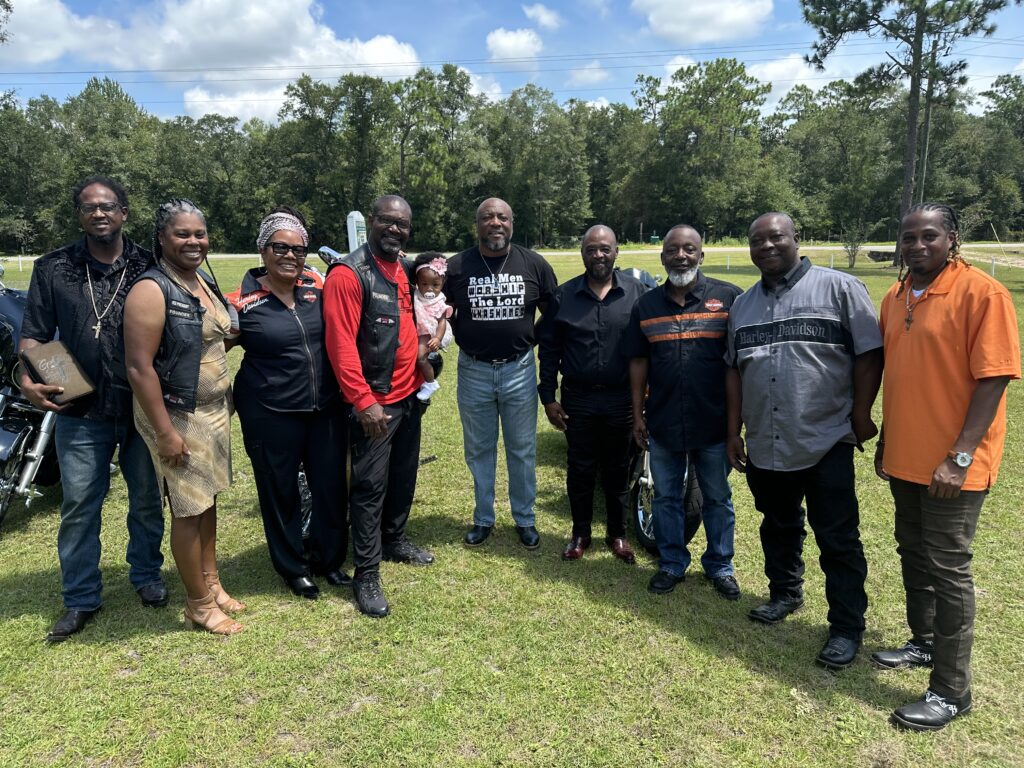 Group of people posing together outdoors on a sunny day, including men and women dressed in motorcycle gear and casual clothing, standing on a grassy lawn with trees and motorcycles in the background.