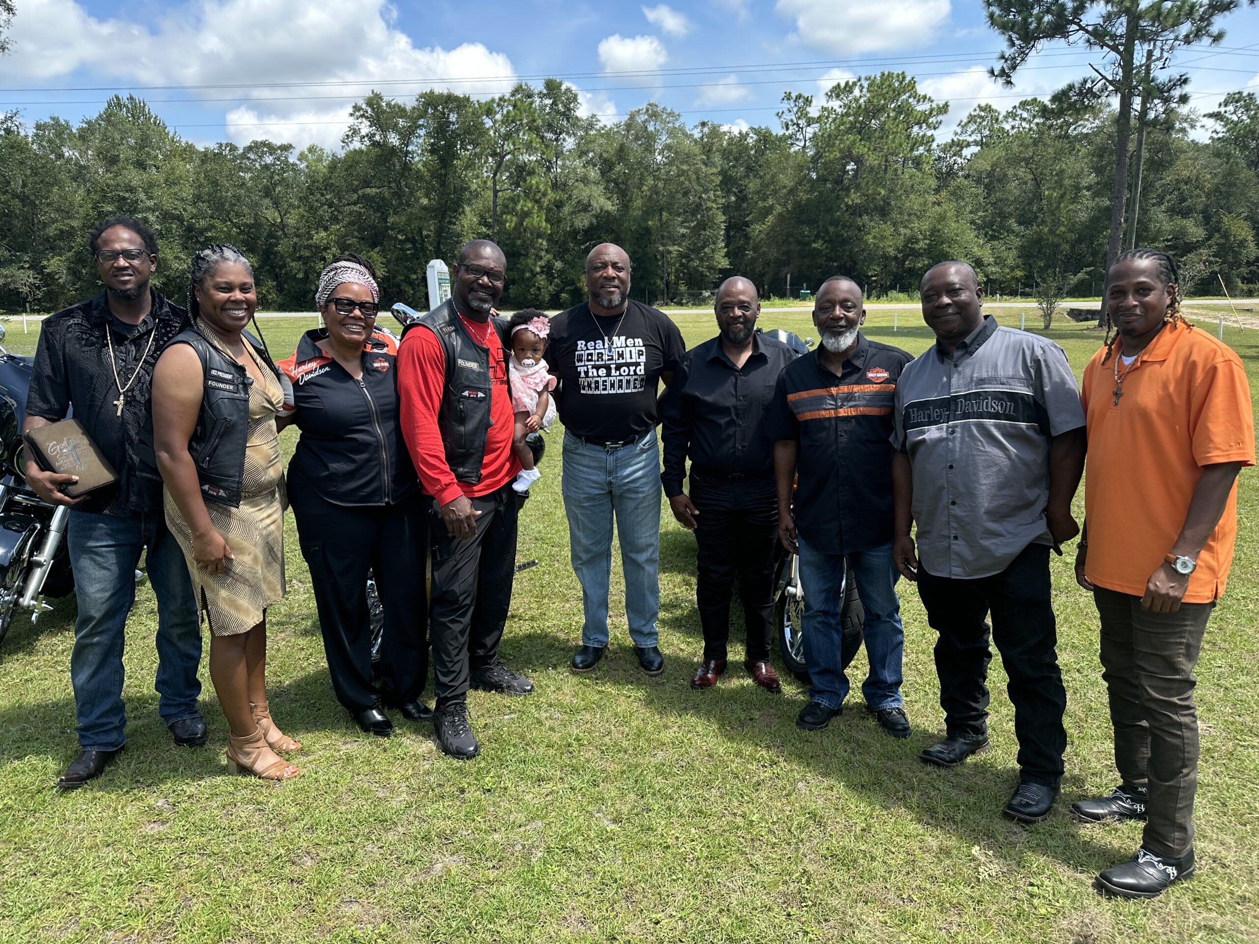Group of people posing together outdoors on a sunny day, including men and women dressed in motorcycle gear and casual clothing, standing on a grassy lawn with trees and motorcycles in the background.