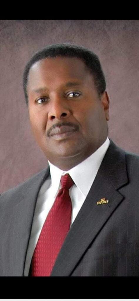 Formal portrait of Reginald James wearing a dark pinstripe suit, white shirt, and red tie, looking confidently at the camera with a neutral background.