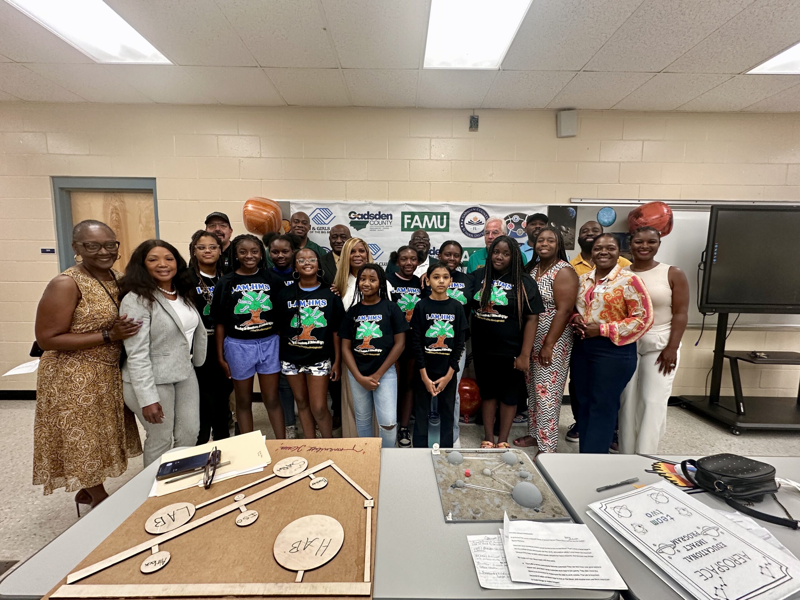 Group photo of students, mentors, and community members standing together in a classroom, with a display of science projects and educational materials on tables in front. A backdrop includes logos for FAMU, Gadsden County, the Boys & Girls Club, and other sponsors.