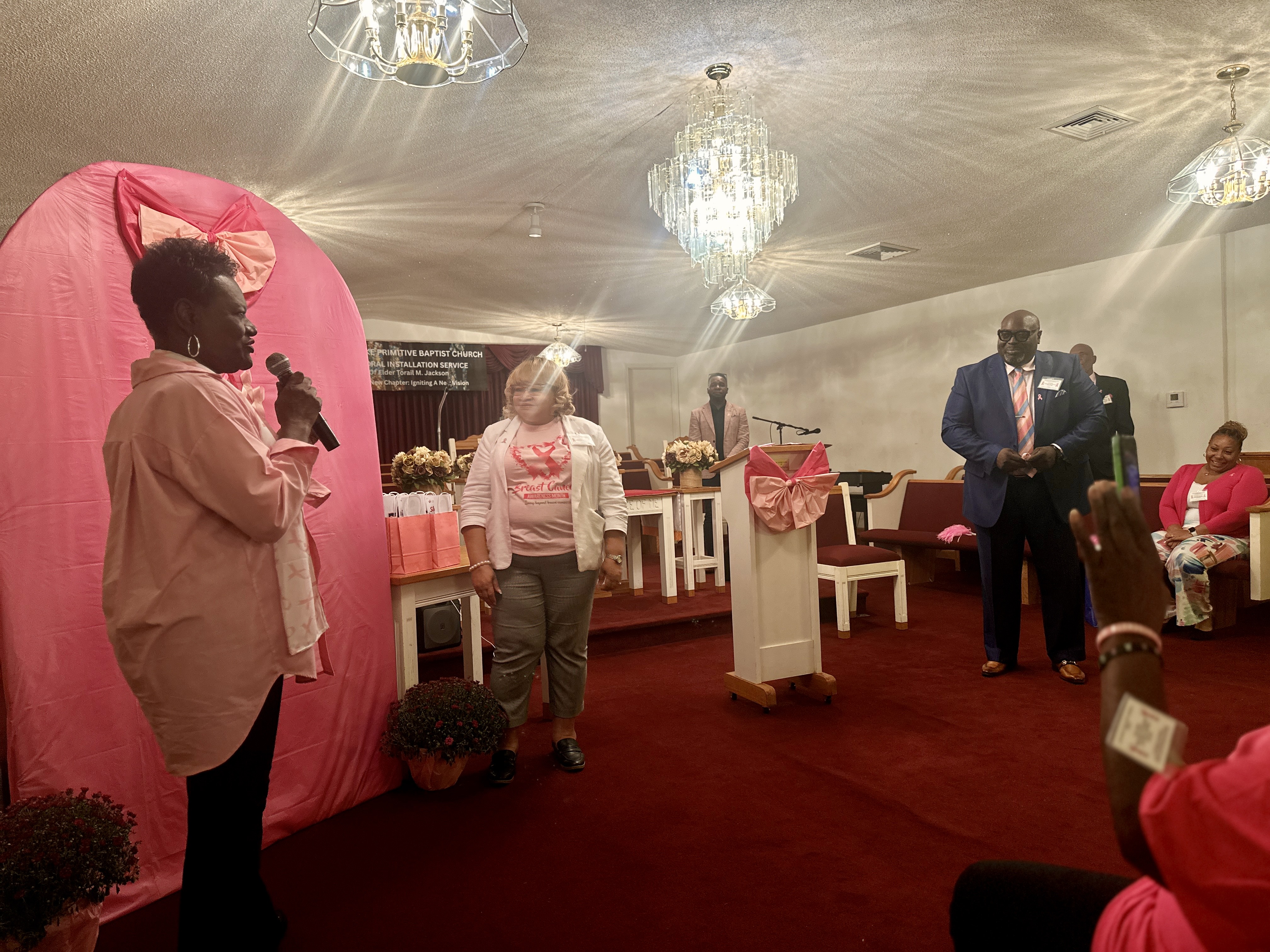 Woman speaking into a microphone in a church decorated with pink ribbons for a breast cancer awareness event, while others look on.