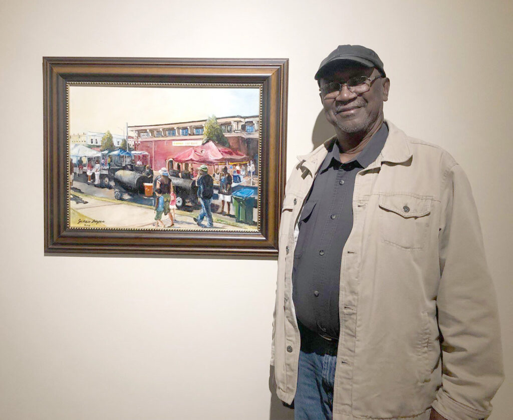Man standing and smiling next to a framed painting depicting an outdoor market scene with tents and people.