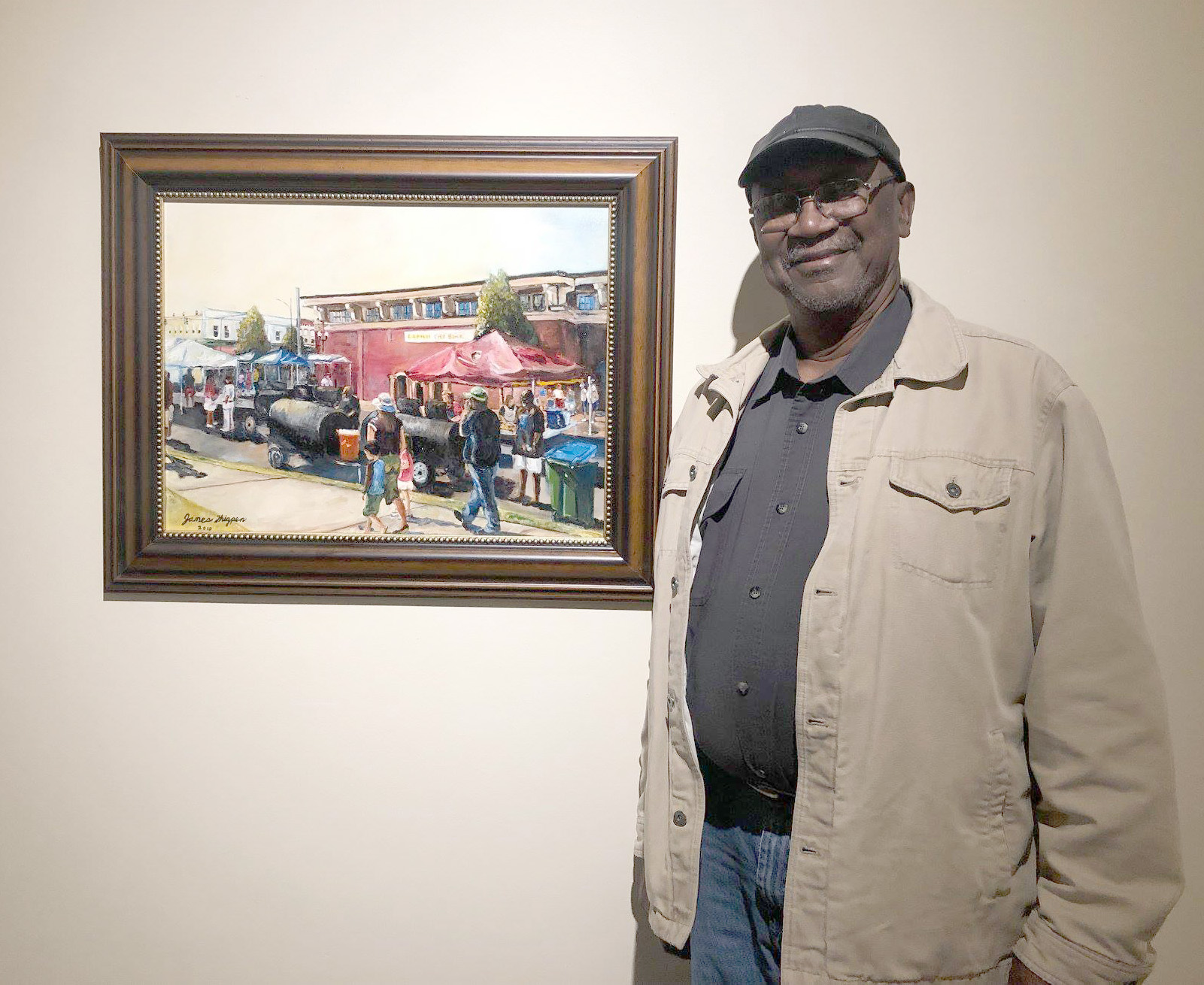Man standing and smiling next to a framed painting depicting an outdoor market scene with tents and people.
