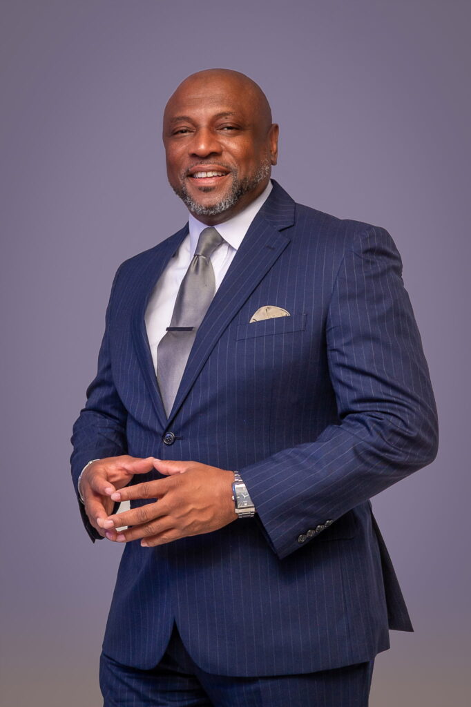 Man in a navy pinstripe suit with a gray tie and pocket square smiling with hands clasped in front against a purple background.