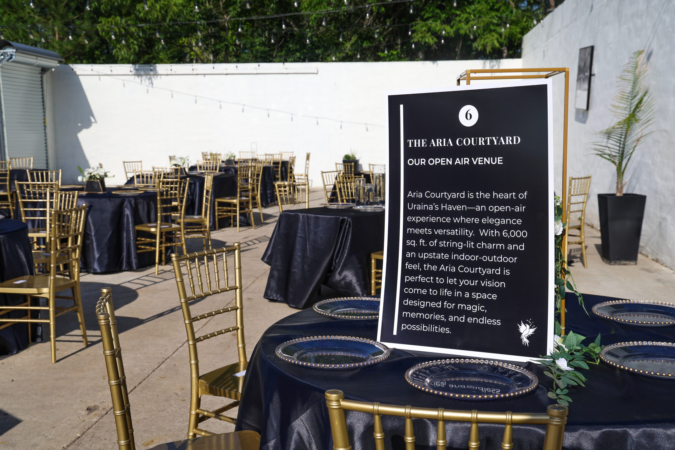 Outdoor courtyard with round tables, gold chairs, and a sign describing “The Aria Courtyard” event venue.
