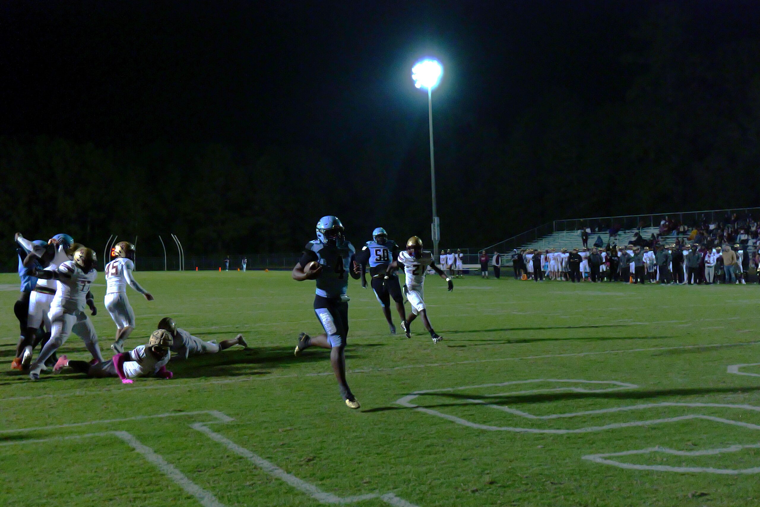 Football player in a blue uniform runs toward the end zone under stadium lights during a night game while defenders chase behind.