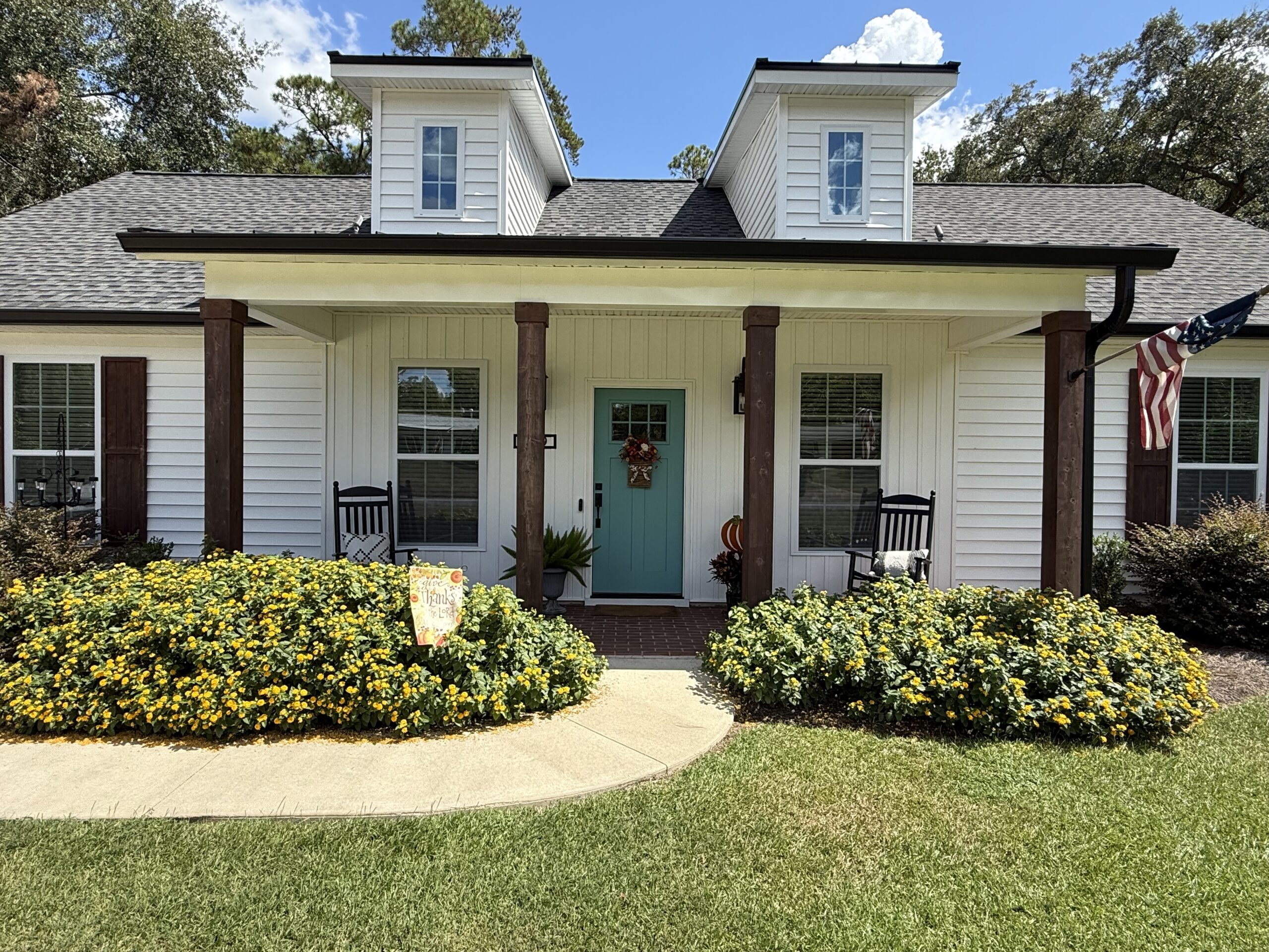 White house with turquoise door, black rocking chairs, and yellow flowers in front yard.