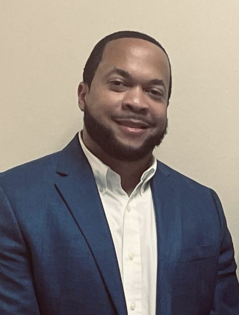 Portrait of a man wearing a blue blazer and white shirt, smiling while standing against a plain indoor background.