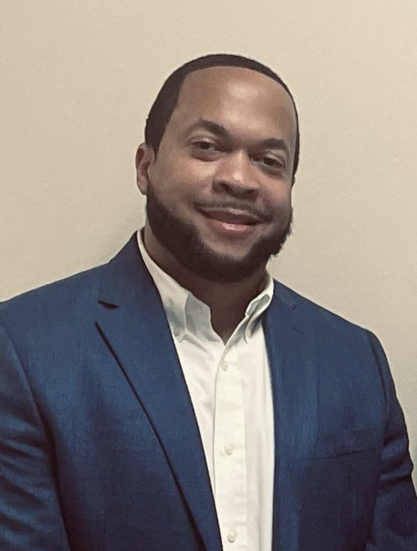 Portrait of a man wearing a blue blazer and white shirt, smiling while standing against a plain indoor background.