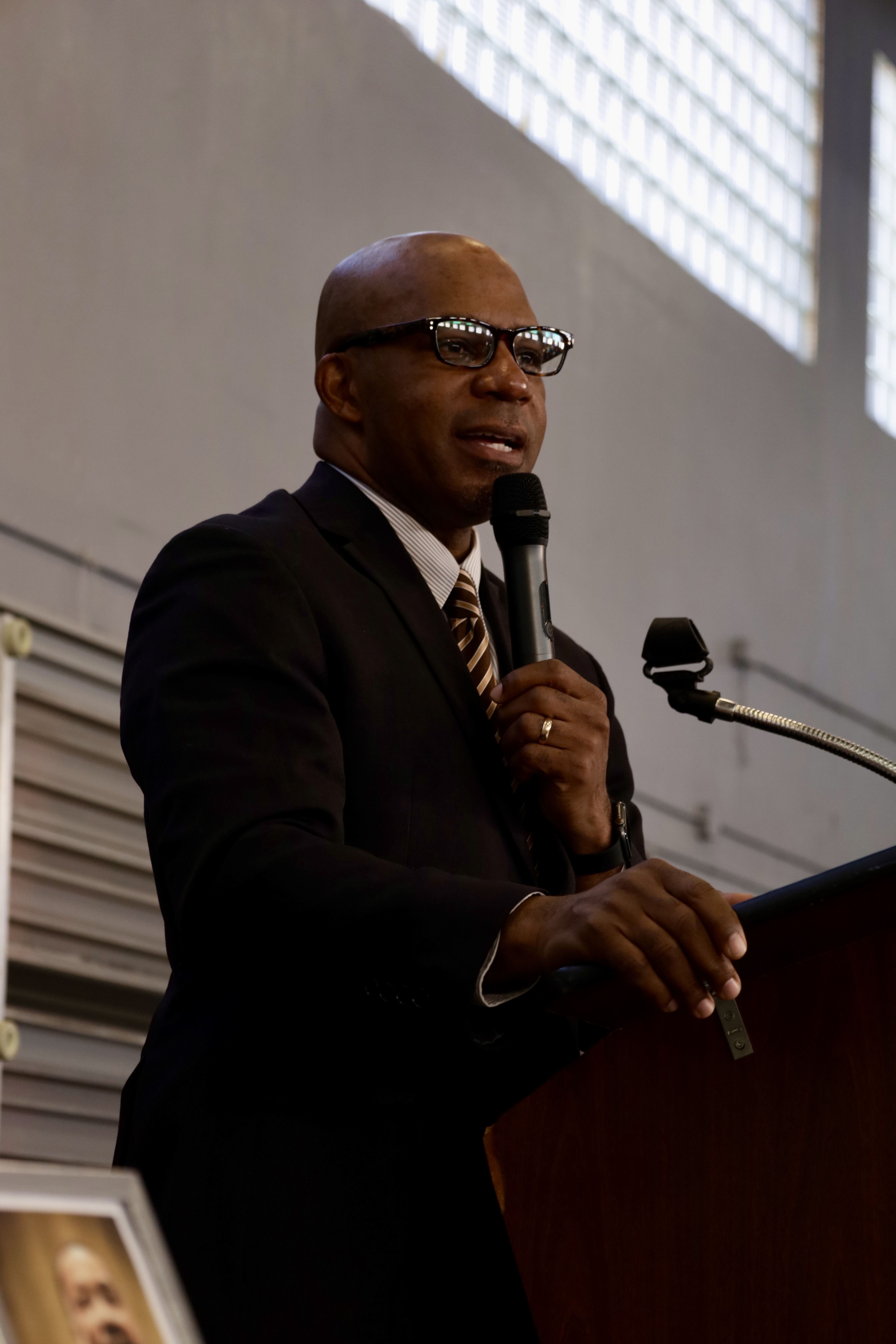 A wide indoor scene showing a group of people standing around round tables inside a gymnasium or community center, appearing attentive during a program or ceremony.