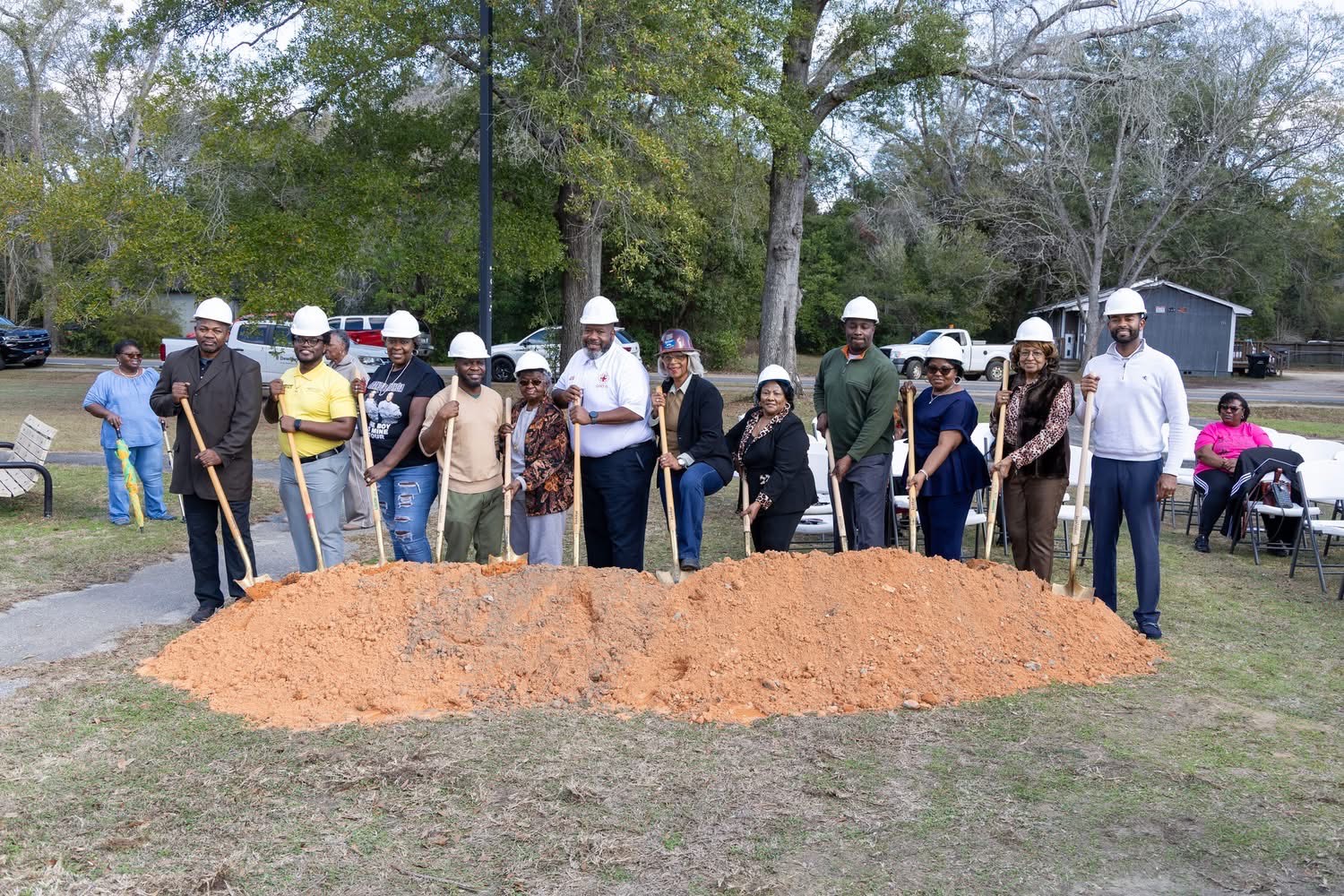 A group of adults wearing white hard hats pose outdoors with ceremonial shovels in front of a mound of dirt, suggesting a groundbreaking ceremony. Trees, parked vehicles, and folding chairs are visible in the background.