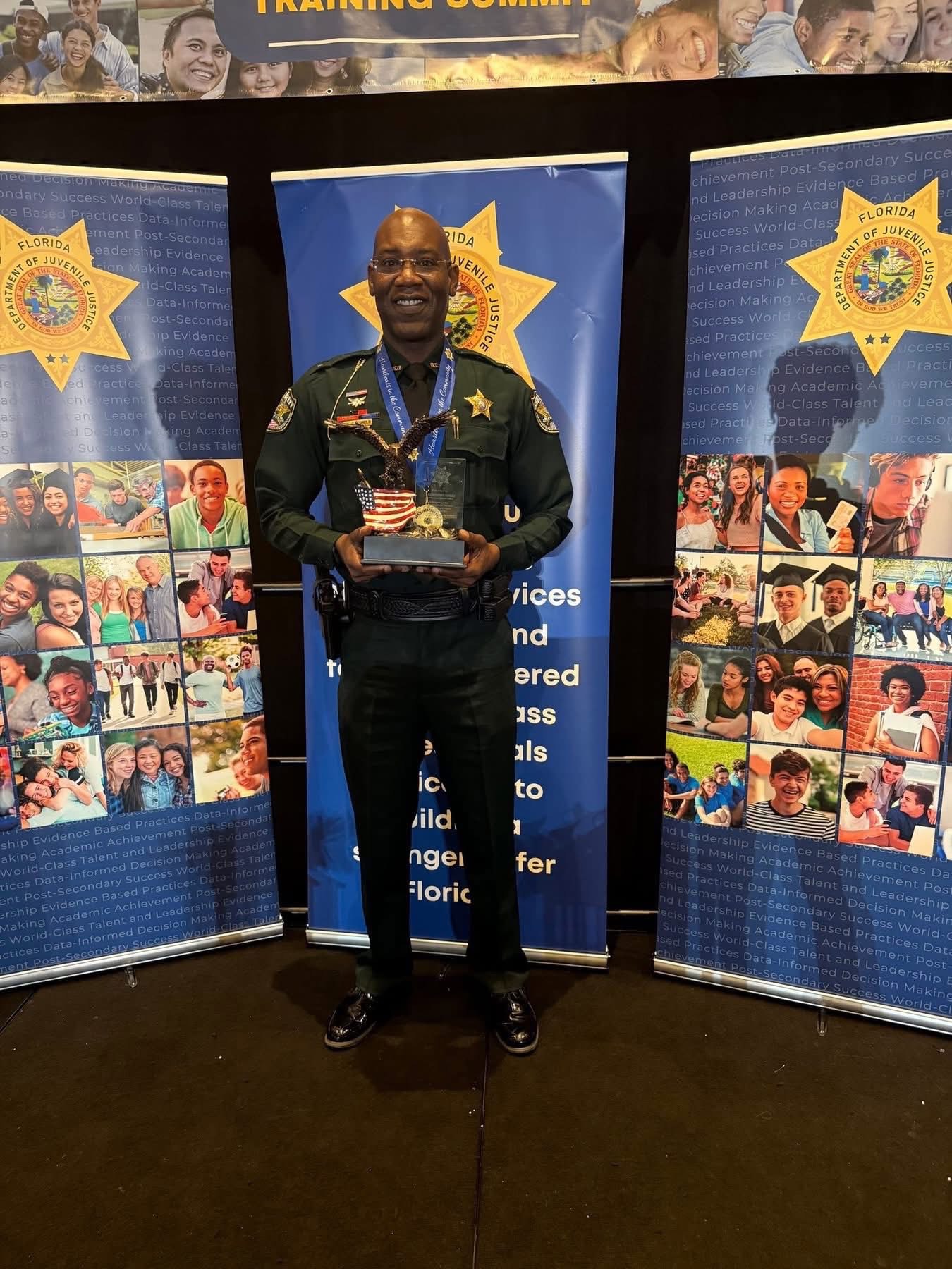 Uniformed law enforcement officer standing indoors between Florida Department of Juvenile Justice banners, holding an eagle statue award mounted on a base with an American flag design.