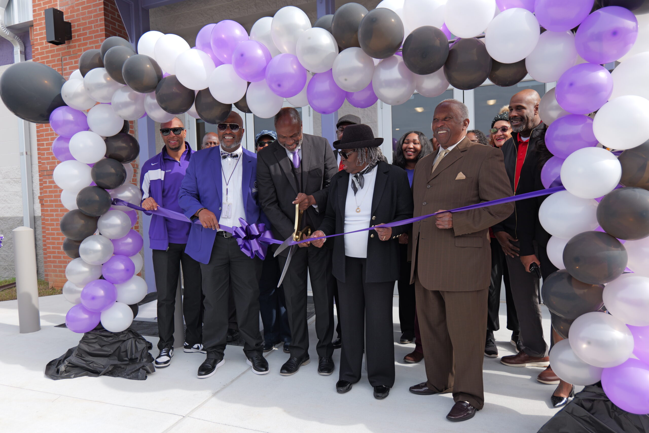 A ribbon-cutting ceremony under a large arch of purple, white, black, and silver balloons. Several adults stand closely together as oversized ceremonial scissors cut a purple ribbon in front of a building entrance.