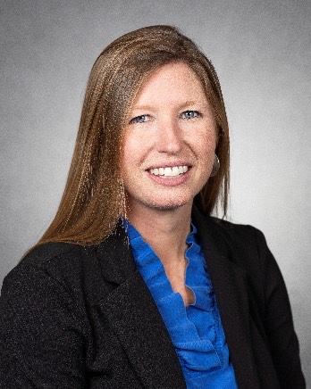 A professional studio-style headshot of a woman wearing a black blazer over a blue blouse. The background is neutral gray, and the subject is smiling directly at the camera.