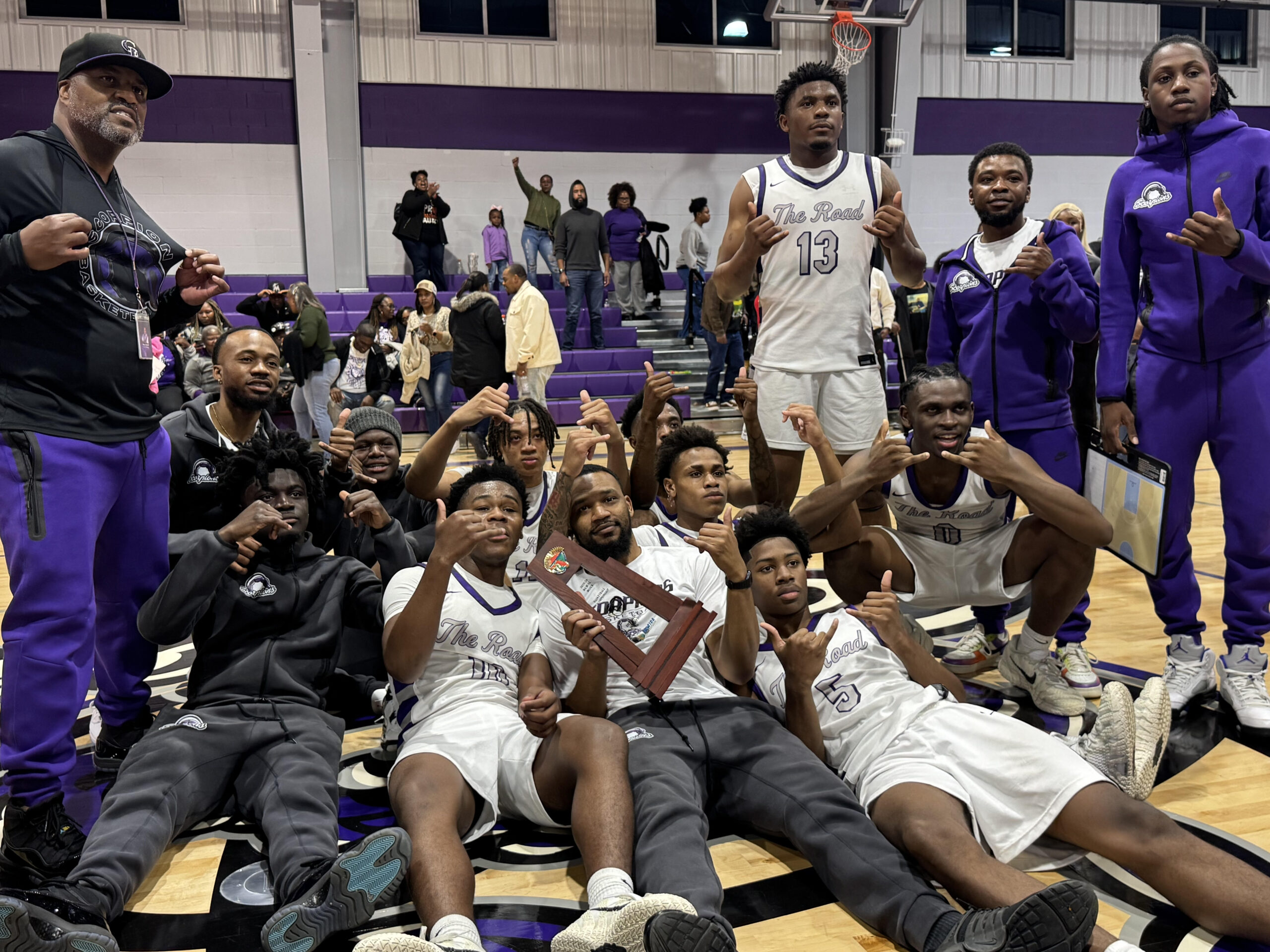 High school basketball players in white uniforms pose on a gym floor with coaches after a game, holding a wooden trophy plaque while fans stand in purple bleachers behind them.