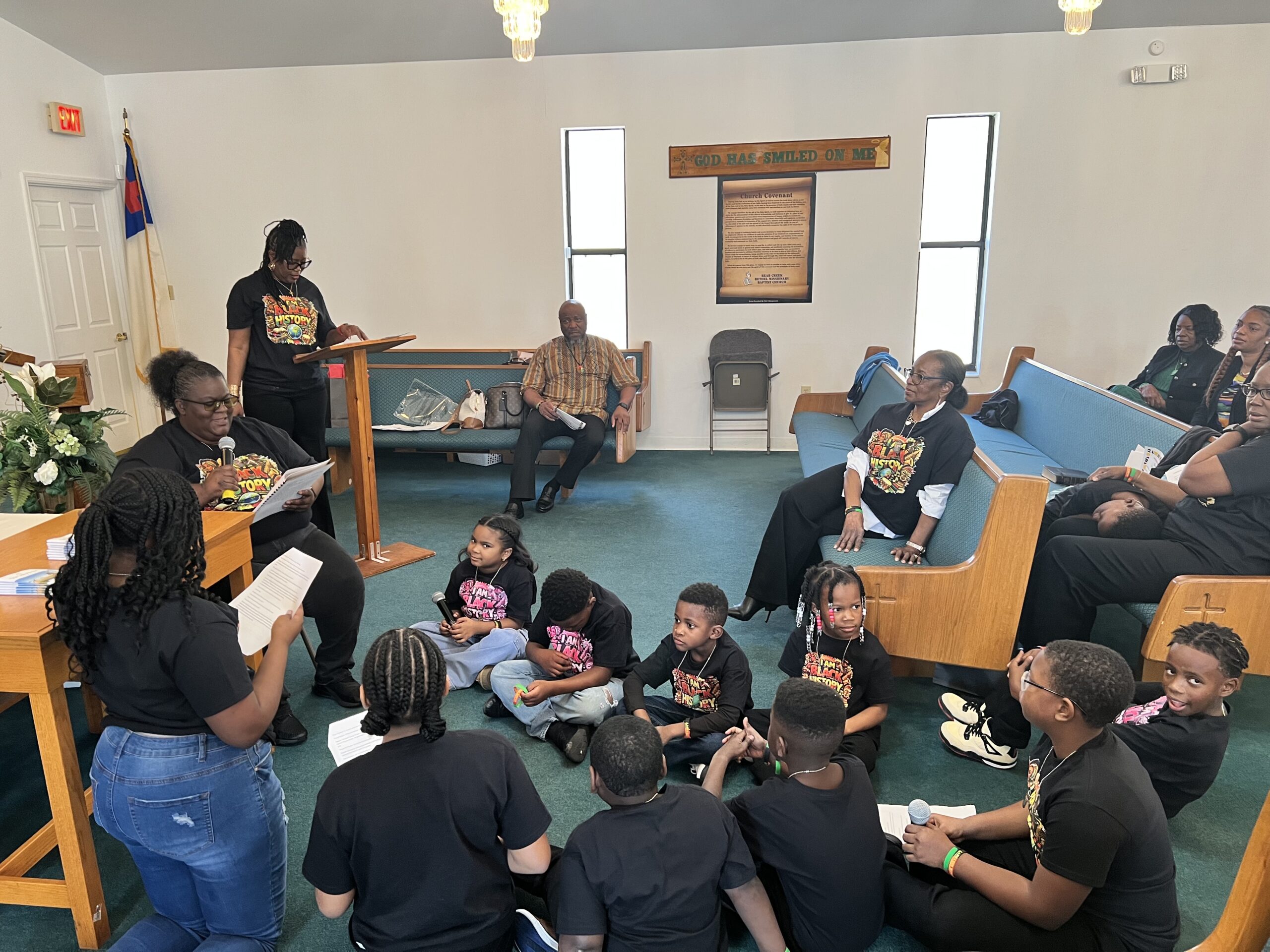 Children and adults wearing matching shirts sit and read together inside a church sanctuary during a group activity.