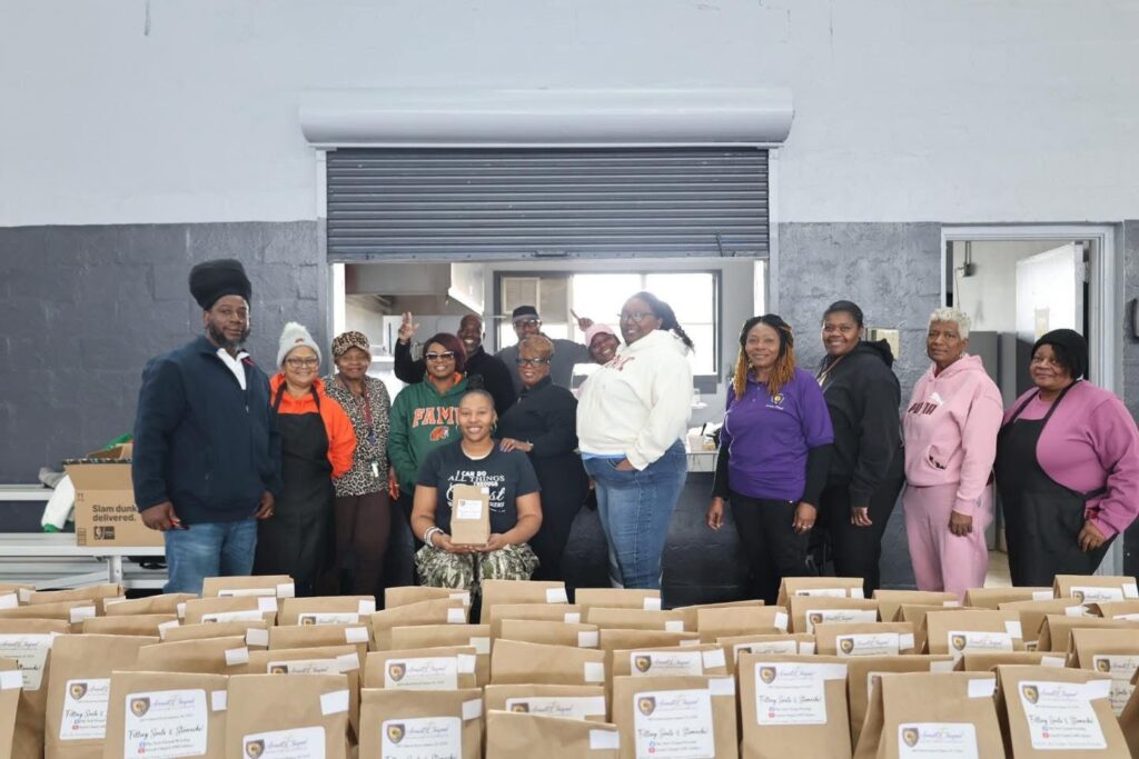 Large group photo indoors showing volunteers standing behind long tables filled with labeled brown paper bags. The group poses and smiles inside a community or gym-like space with a roll-up door in the background.