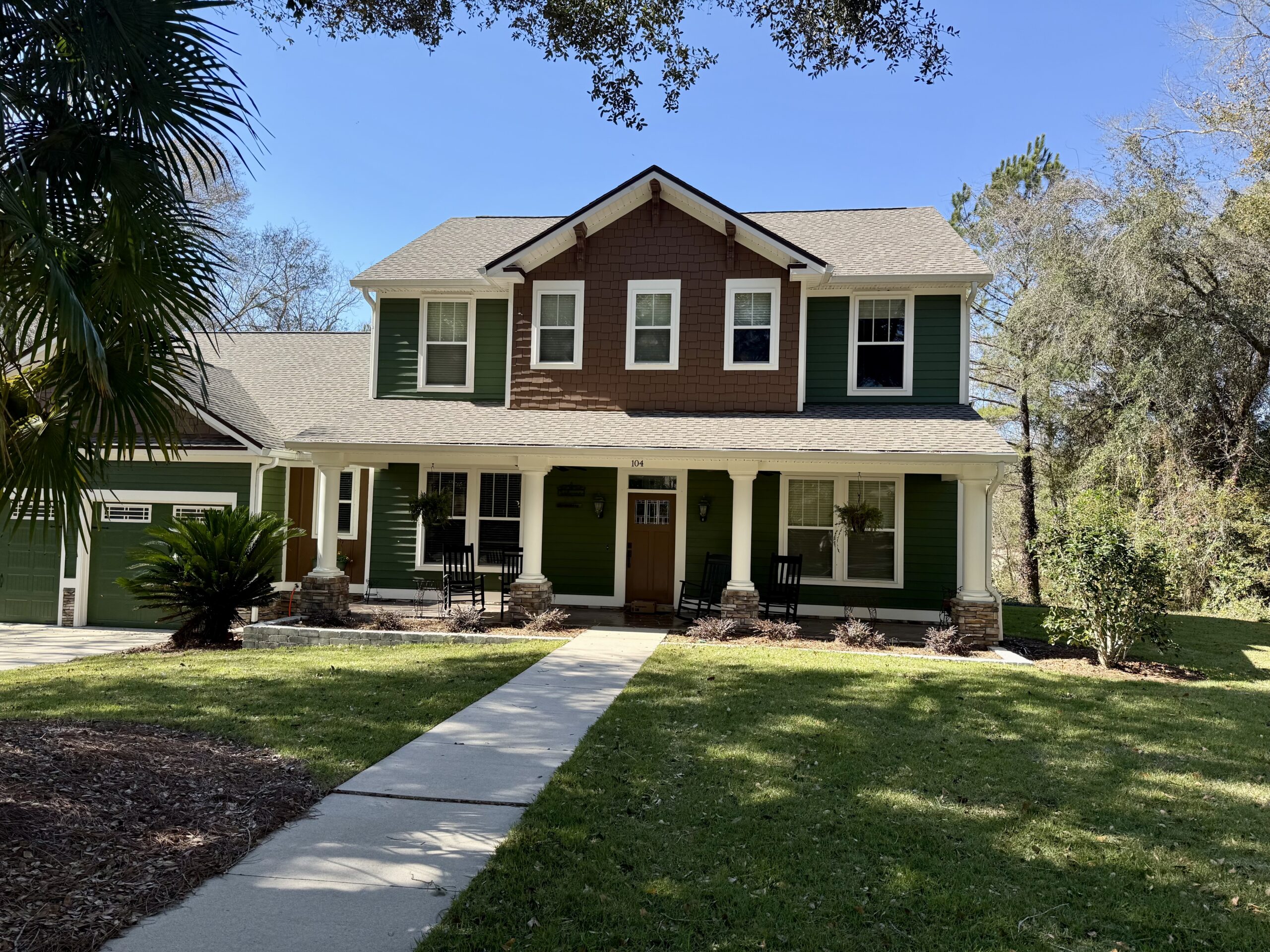 A two-story green house with white columns and a covered front porch sits behind a neatly maintained lawn and walkway.