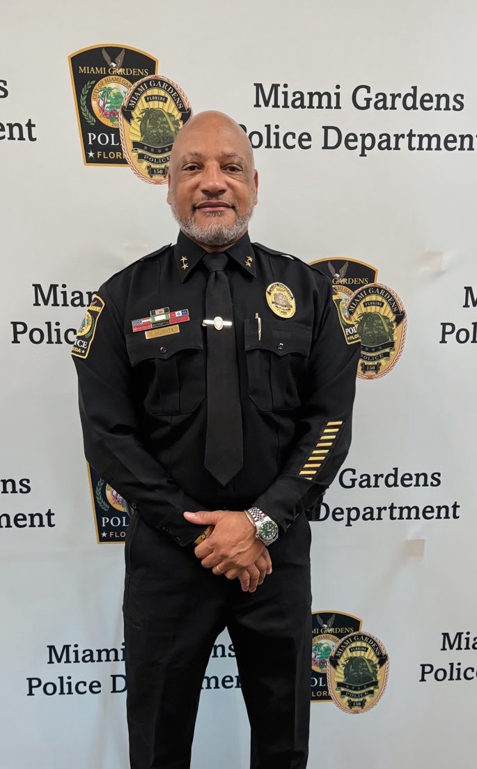 Portrait of a Miami Gardens Police Department officer in full uniform standing in front of a department backdrop displaying official insignia.