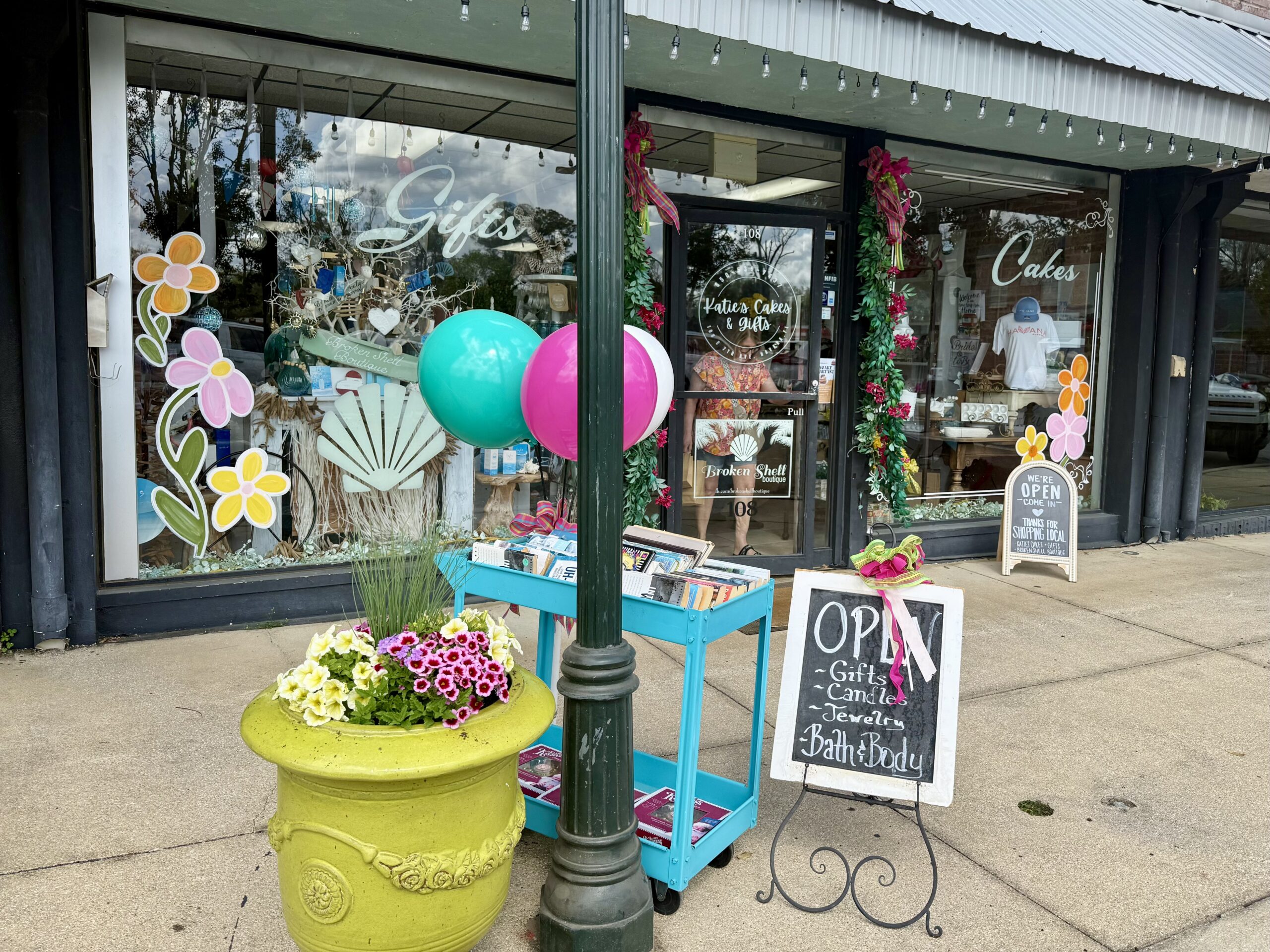 Storefront of Katie’s Cakes & Gifts boutique decorated with balloons, flowers, and window displays advertising gifts and cakes along a downtown sidewalk.