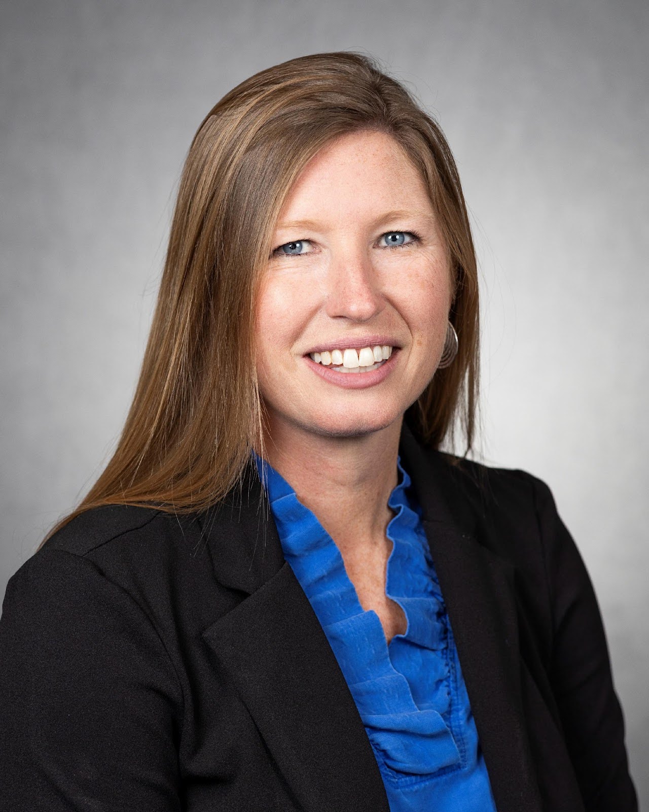 Professional headshot of a woman with long auburn hair wearing a black blazer and blue blouse, smiling against a neutral studio background.