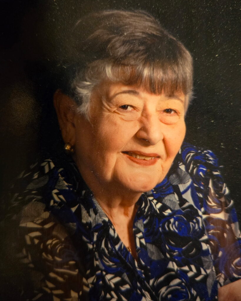 Portrait of an elderly woman with short gray hair wearing a patterned blouse, smiling against a dark studio background.