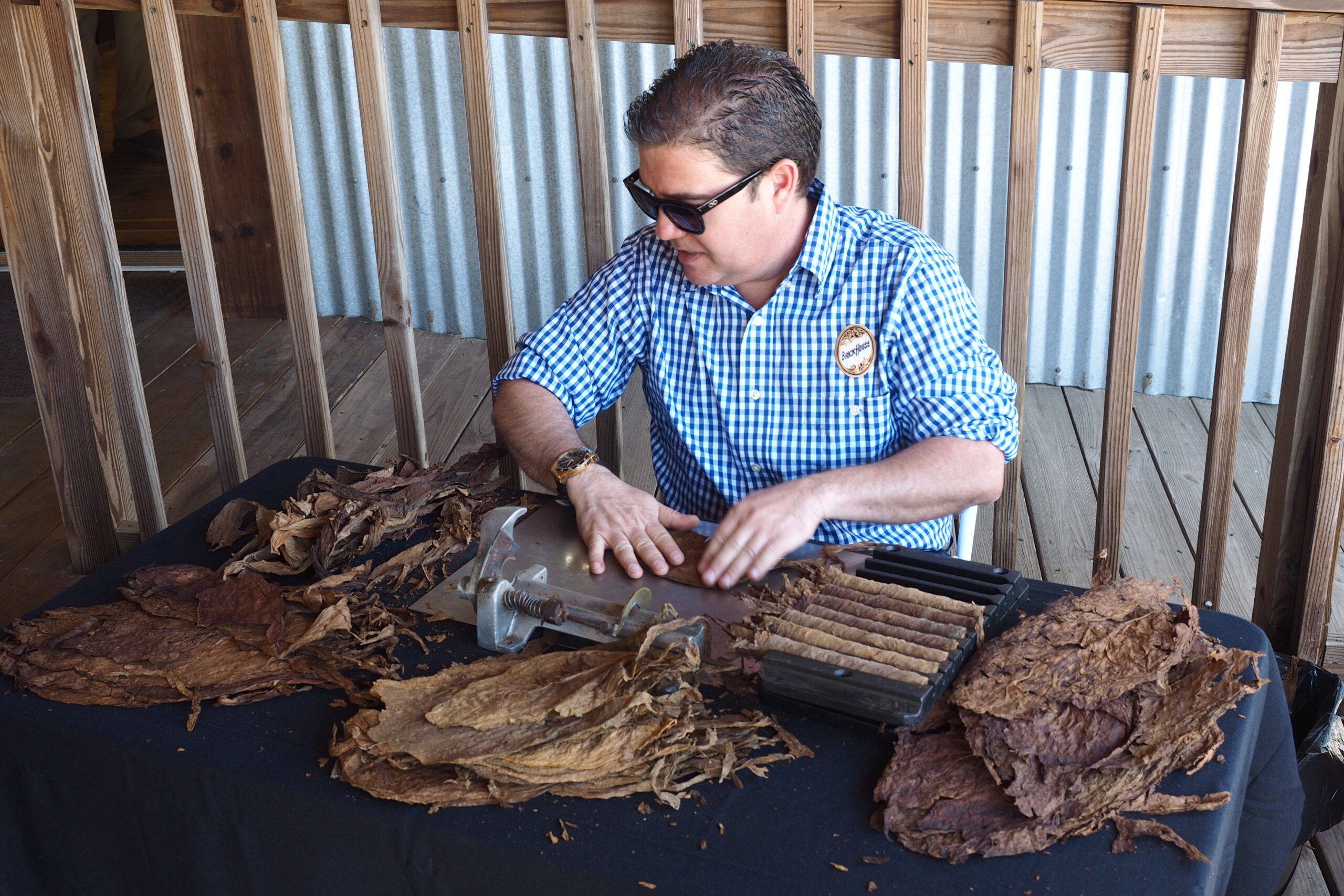 Man identified as cigar roller Lois Gonzalez shaping tobacco leaves on a rolling table, with stacks of dried tobacco and finished cigars arranged around him.