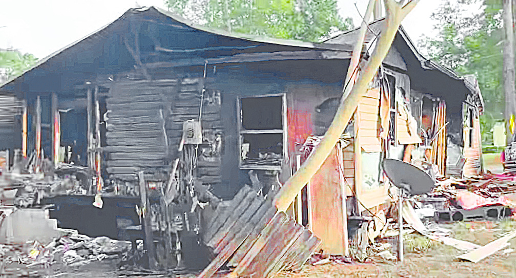 Severely burned house with heavy fire damage, charred siding, and debris scattered around the structure.