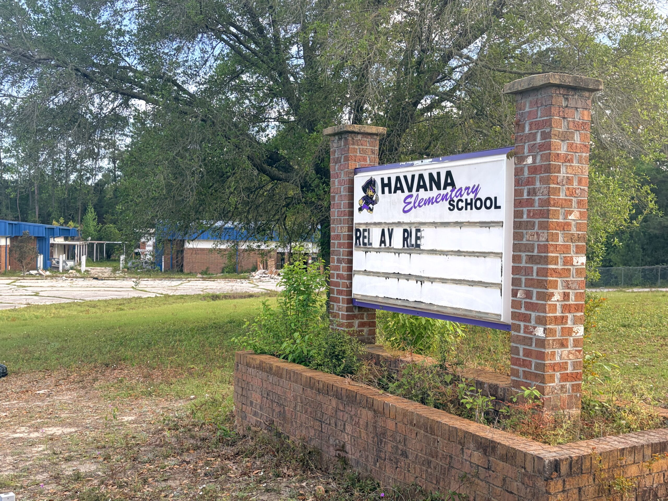 Sign for Havana Elementary School in front of an older campus with overgrown grass and buildings in the background, with the message board partially reading “RELAY RE.”