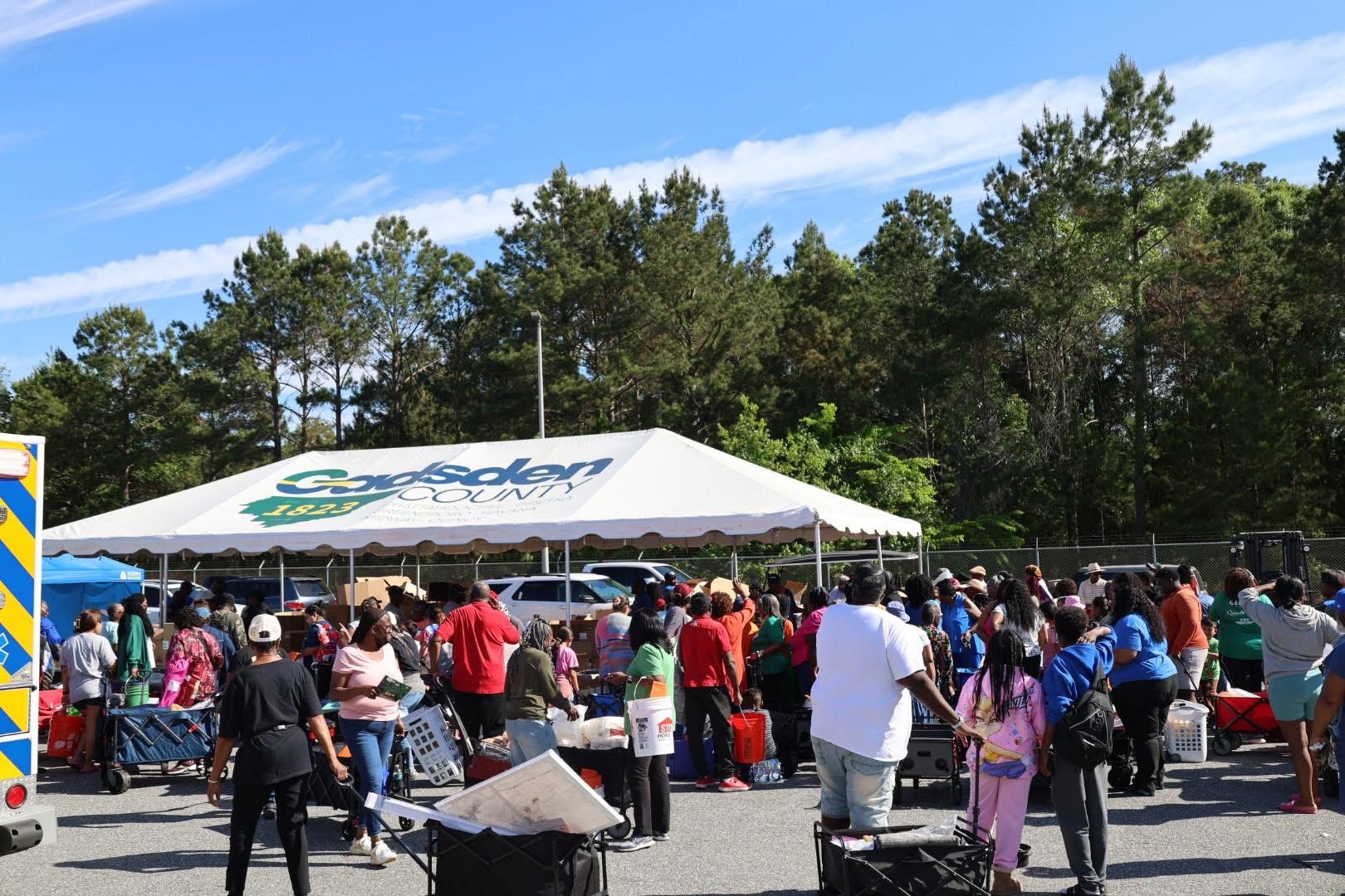 Large crowd gathered around a tent labeled Gadsden County, with people collecting items and pushing carts during a community distribution event.