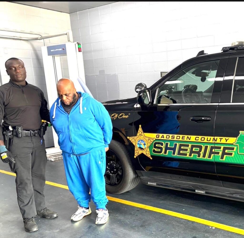 Man in a blue tracksuit standing in handcuffs next to a Gadsden County Sheriff’s patrol vehicle while a deputy stands nearby.