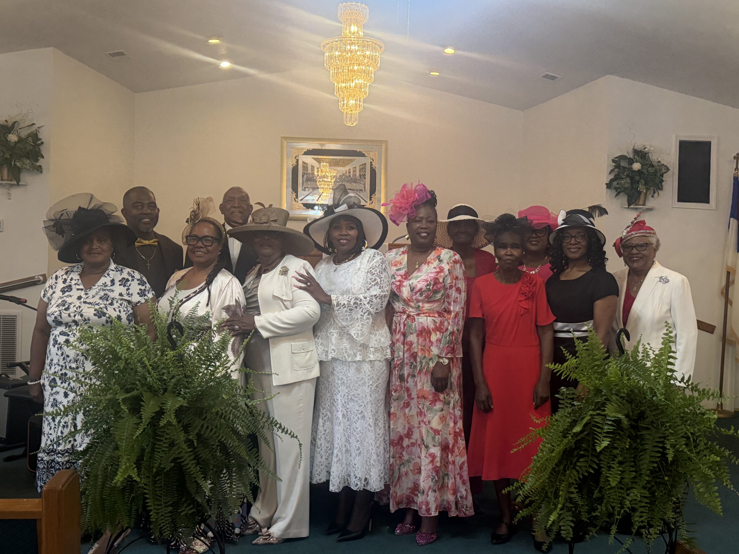 A group photo from the Pretty Hats & Bow Ties event at Bear Creek Bethel Missionary Baptist Church. A group of Black congregants, primarily women wearing elegant, colorful dresses and elaborate, wide-brimmed church hats or fascinators, pose together at the front of the sanctuary. Two men wearing bow ties stand in the background, and large green ferns frame the group in the foreground under a glowing crystal chandelier