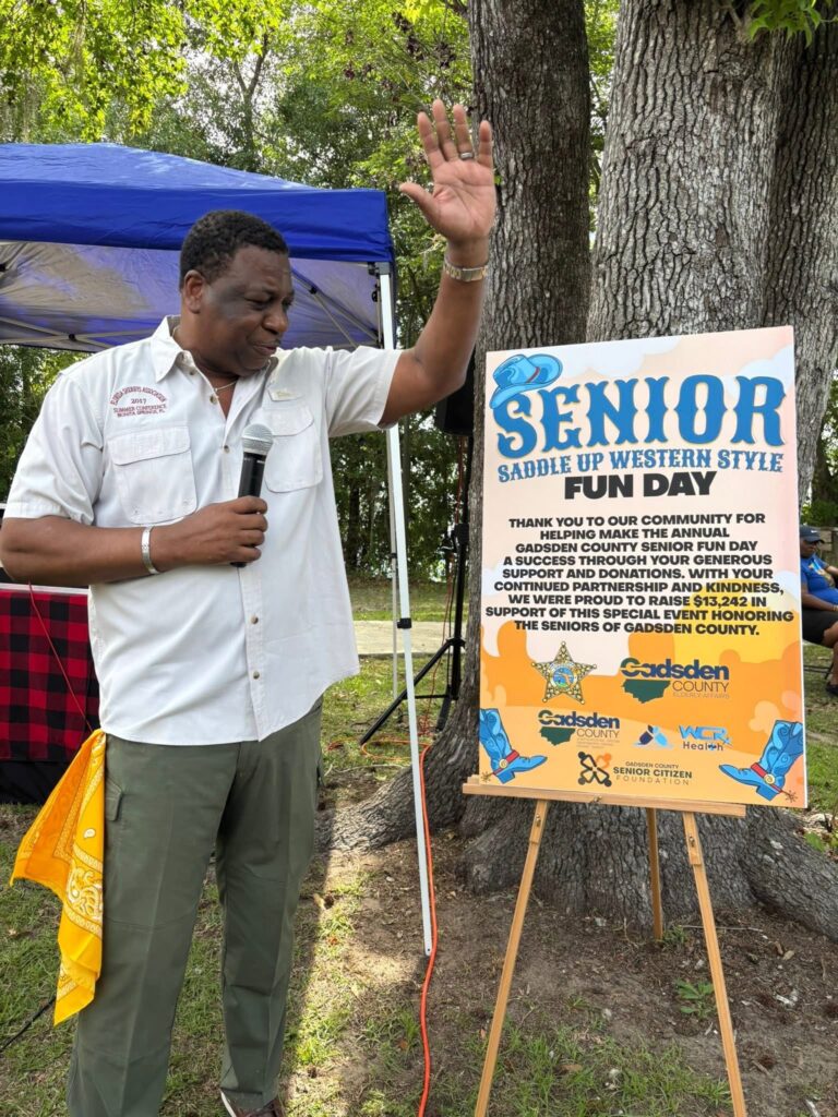 A man holding a microphone raises his hand beside a sign for the Senior Saddle Up Western Style Fun Day event outdoors beneath large trees.