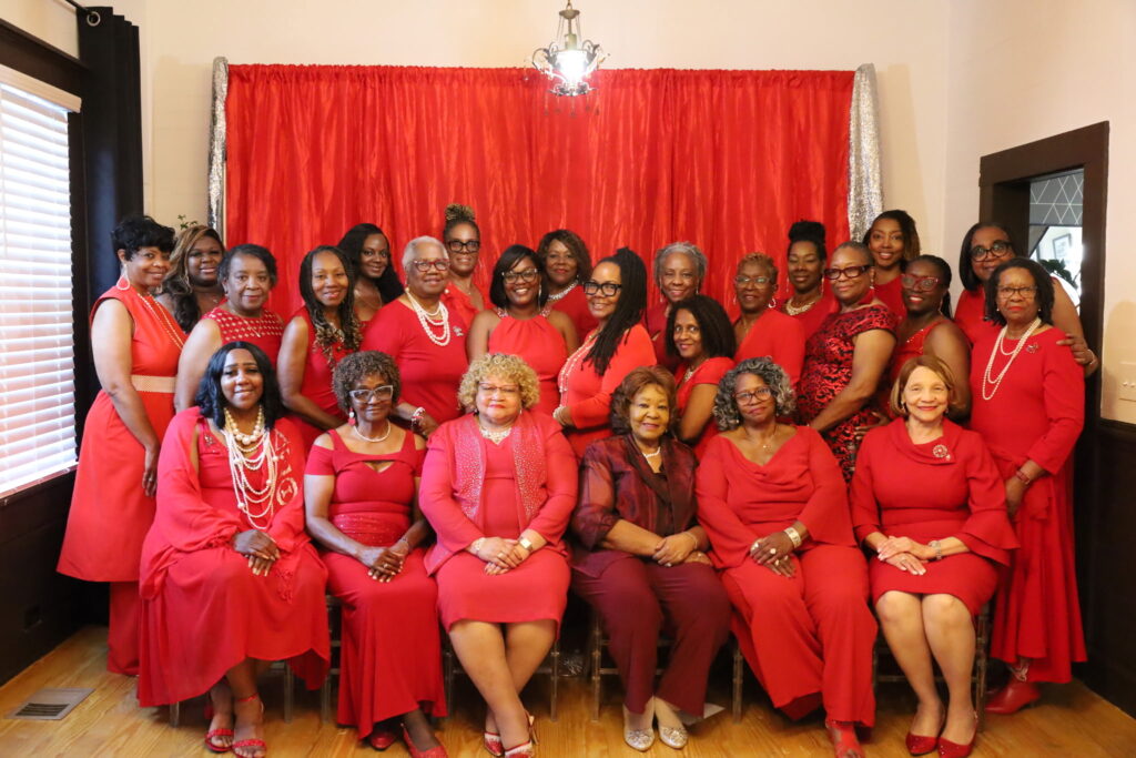 A large group photograph of more than twenty women dressed entirely in shades of red, burgundy, and pink posing together indoors. The group features a front row of seated women and two standing rows behind them, with many wearing pearl necklaces and jewelry. They are positioned in front of a bright red fabric backdrop framed by silver tinsel curtains.