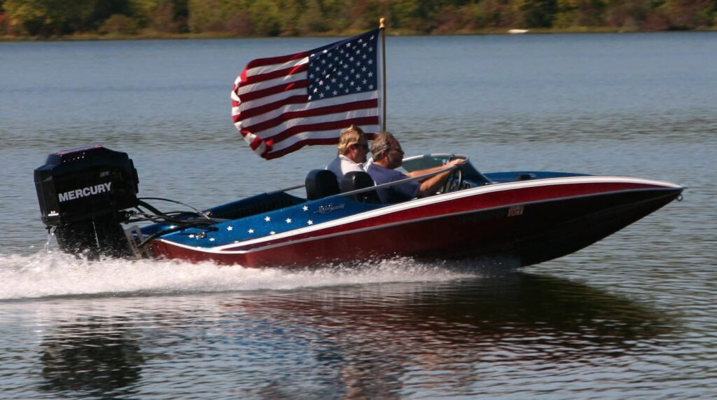 A red, white, and blue patriotic-themed speed boat cruising fast on the water for the Chattahoochee Thunder Nationals on the Apalachicola River. Two men are inside the boat, which features white stars on its blue trim, a large black Mercury outboard motor on the back, and a large American flag waving from a flagpole behind the seats.