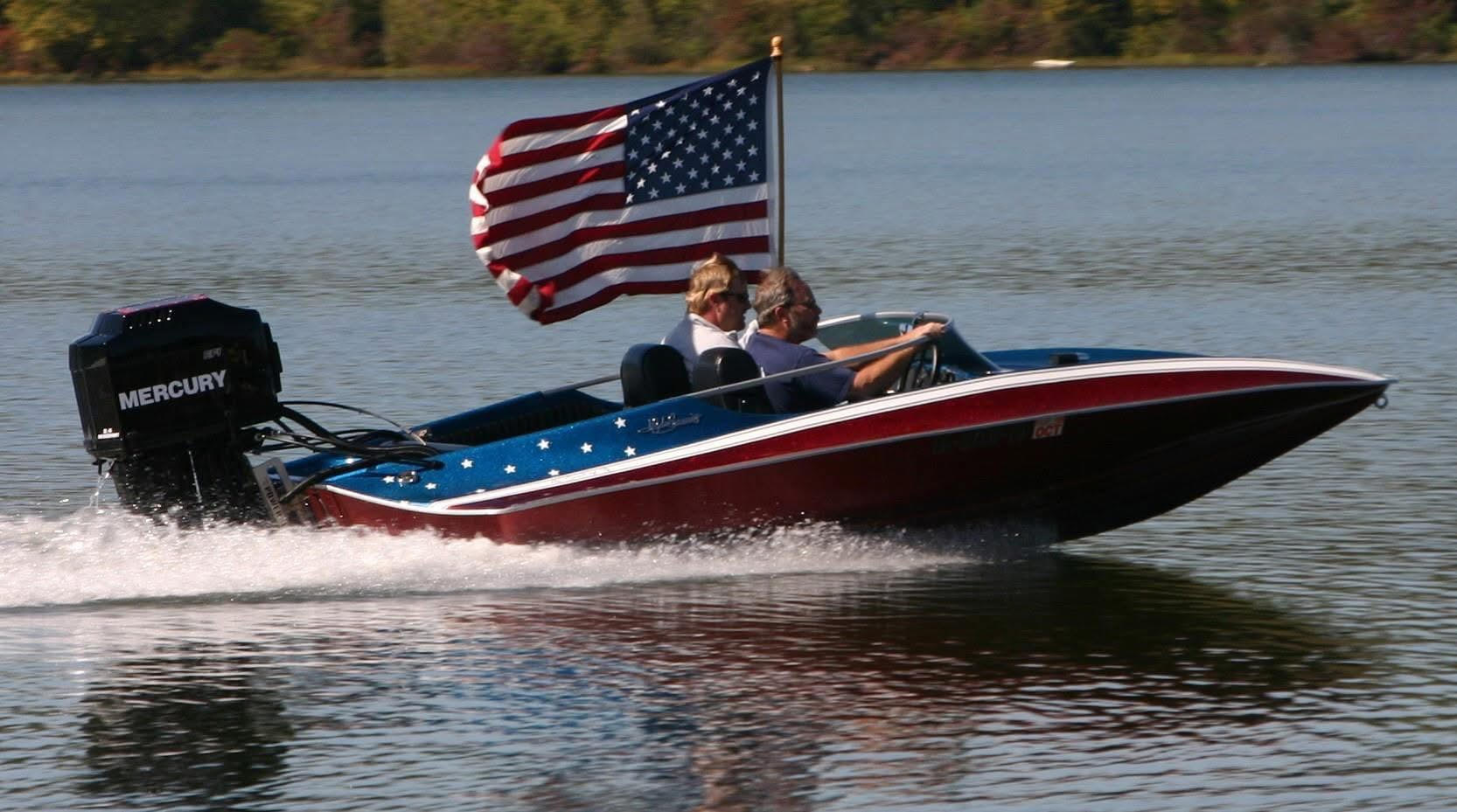 A red, white, and blue patriotic-themed speed boat cruising fast on the water for the Chattahoochee Thunder Nationals on the Apalachicola River. Two men are inside the boat, which features white stars on its blue trim, a large black Mercury outboard motor on the back, and a large American flag waving from a flagpole behind the seats.
