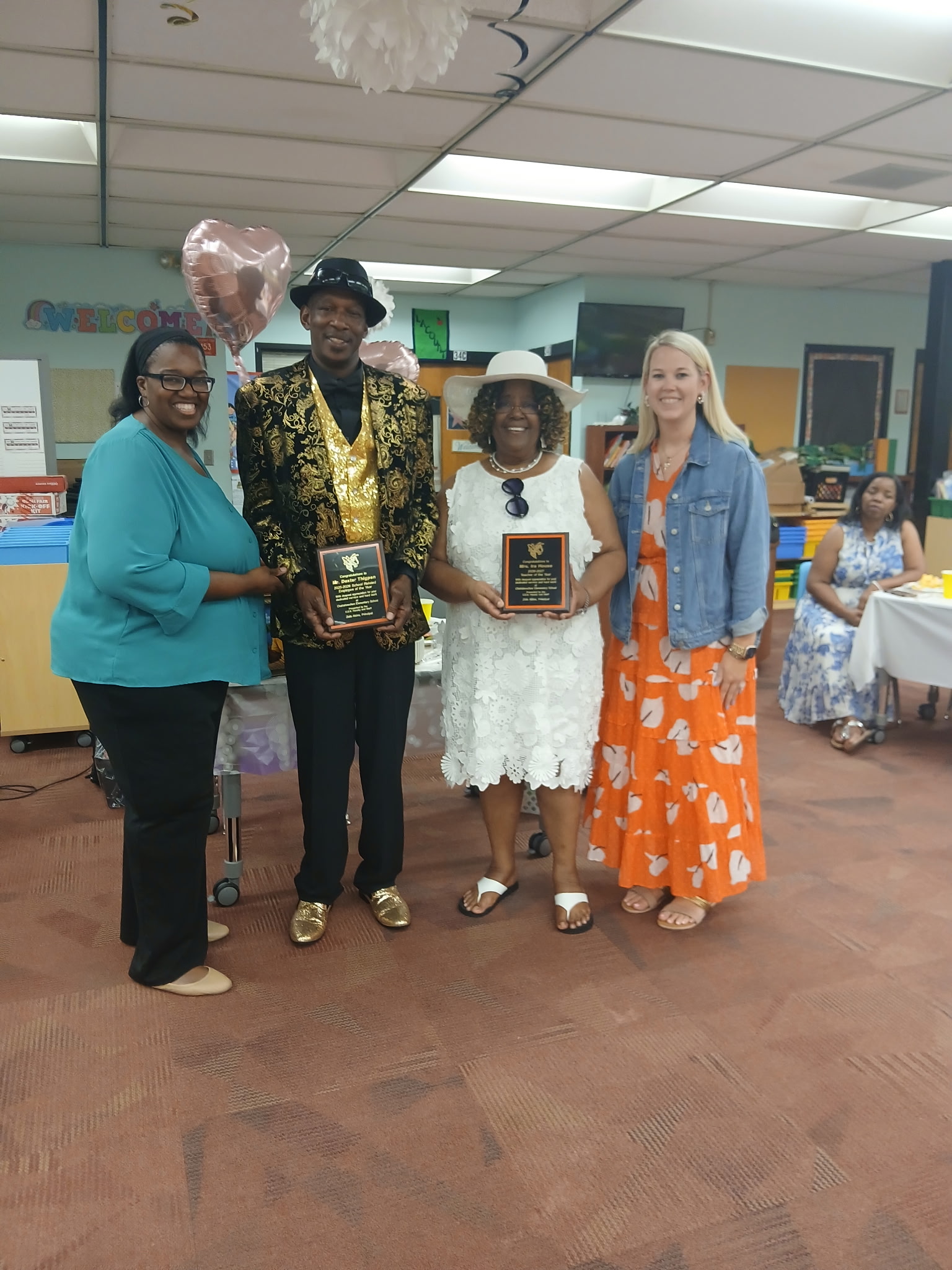 An awards presentation inside a school classroom, featuring four adults posing together in the foreground. A man dressed in a black hat, a black-and-gold patterned blazer, a gold sequined vest, and gold shoes stands next to a woman wearing a white lace dress and a white sun hat; both are holding black recognition plaques. They are flanked by two smiling women, one on the left in a teal blouse and black pants, and one on the right in an orange patterned dress and denim jacket.