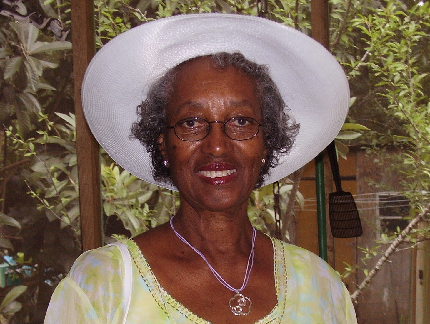Obituary portrait of Elma Cummings. The photograph shows an older Black woman smiling warmly at the camera, wearing an elegant cream-colored dress or jacket and a matching wide-brimmed church hat with lace detailing against a neutral studio background.