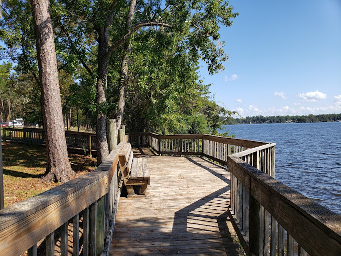 A wooden boardwalk with handrails overlooks a wide, calm lake on a clear, sunny day. A wooden park bench is positioned along the walkway under the shade of mature trees lining the grassy shoreline. Shadows stretch across the wooden planks, and small white clouds dot the blue sky over the distant opposite shore.