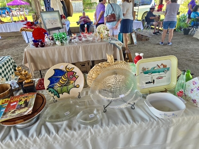 A close-up view of a vendor table. The table is covered in a white tablecloth and displays various collectible items, including fish-shaped glass plates, a colorful ceramic fish platter, a vintage metal tray featuring an illustration of a classic car, old glassware, and a retro 'Jack and Jill' children's magazine. In the background, other shoppers browse additional tables under the shaded pavilion.