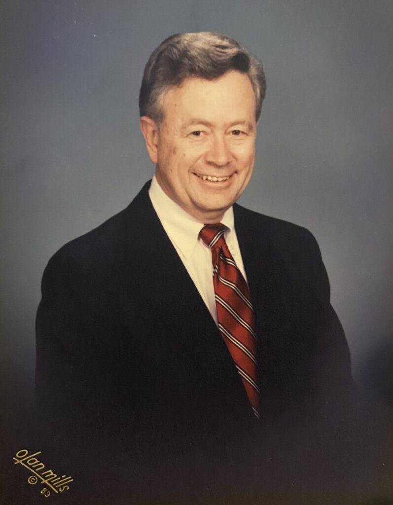 A professional studio headshot portrait of an older man smiling warmly. He has short, styled graying hair and is wearing a dark suit jacket, a white collared dress shirt, and a red tie with dark diagonal stripes. The photograph is shot from the chest up against a mottled blue-gray studio background, with an 'Olan Mills © 89' signature visible in the bottom-left corner.