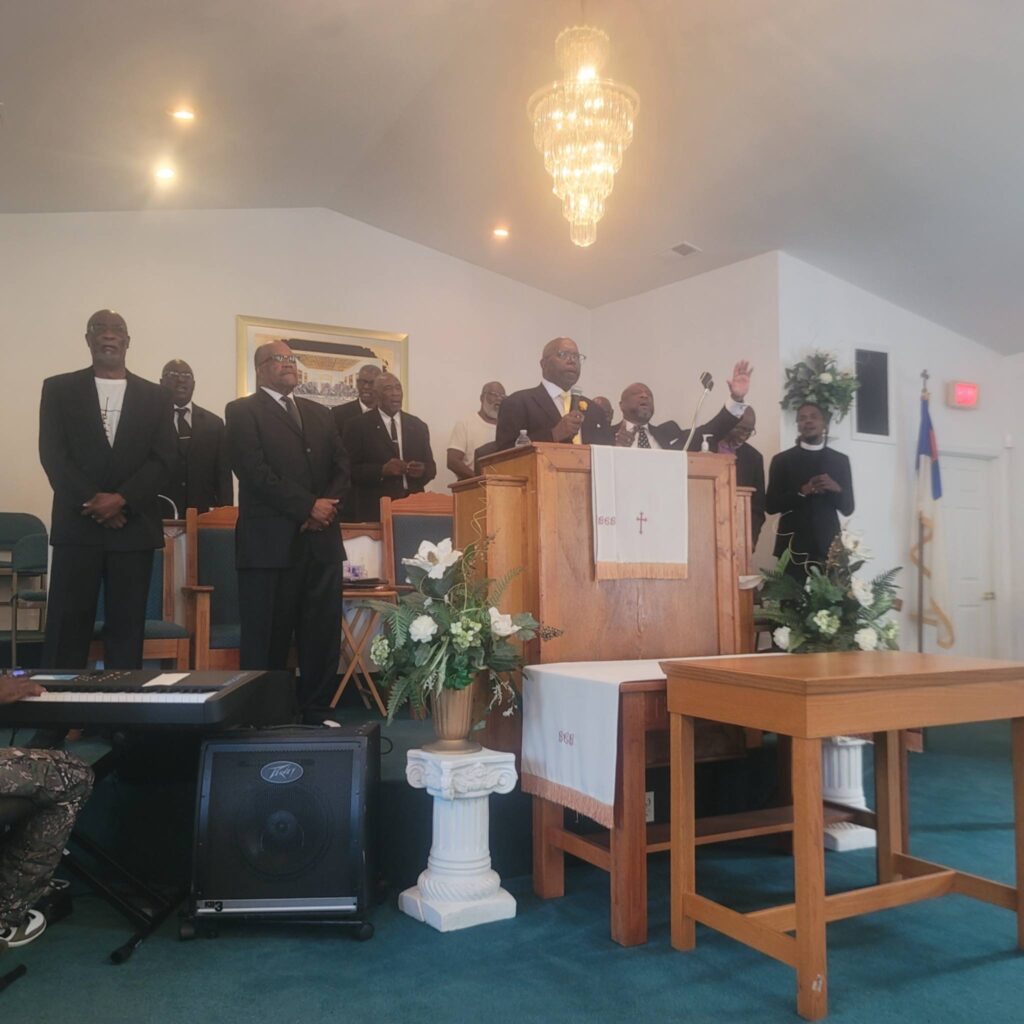 Reverend Danny Whitfield preaches from a central wooden pulpit with one hand raised at Bear Creek Bethel Missionary Baptist Church during an 'Iron Sharpens Iron' event. He is surrounded by several men in dark suits standing on the stage, including Pastor Terrence D. Milton to the left of the pulpit. The sanctuary stage features white floral arrangements, an altar table, a crystal chandelier hanging above, and musical equipment in the foreground.