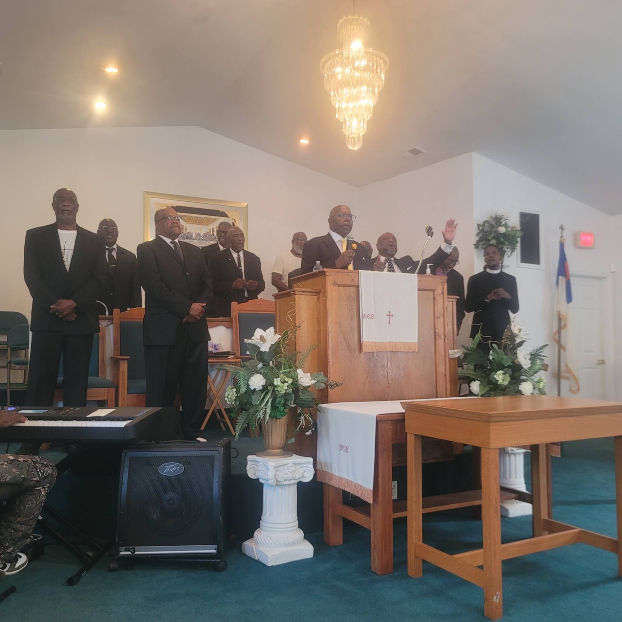Reverend Danny Whitfield preaches from a central wooden pulpit with one hand raised at Bear Creek Bethel Missionary Baptist Church during an 'Iron Sharpens Iron' event. He is surrounded by several men in dark suits standing on the stage, including Pastor Terrence D. Milton to the left of the pulpit. The sanctuary stage features white floral arrangements, an altar table, a crystal chandelier hanging above, and musical equipment in the foreground.