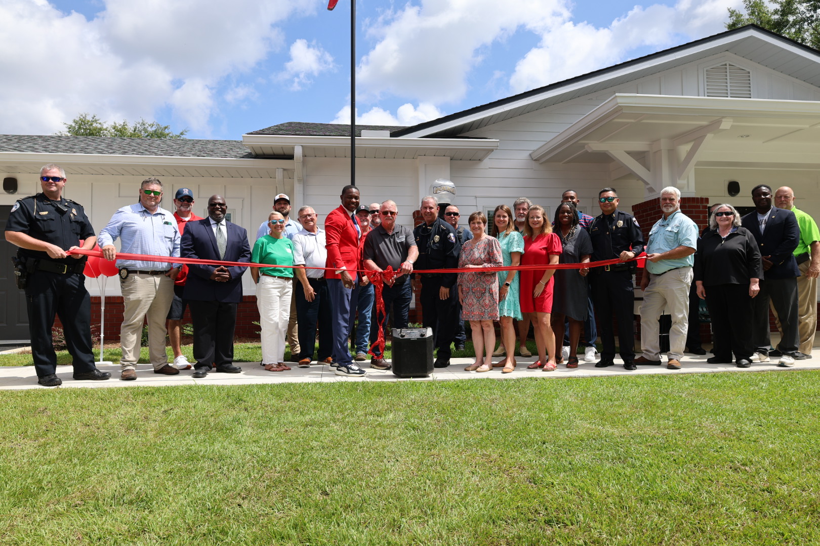 A large group of local officials, police officers, and community members gathered outside a white single-story building for a ribbon-cutting ceremony under a blue sky. Two police officers hold a long red ribbon across the front lawn while a man in a red suit jacket stands near the center next to others holding giant scissors.