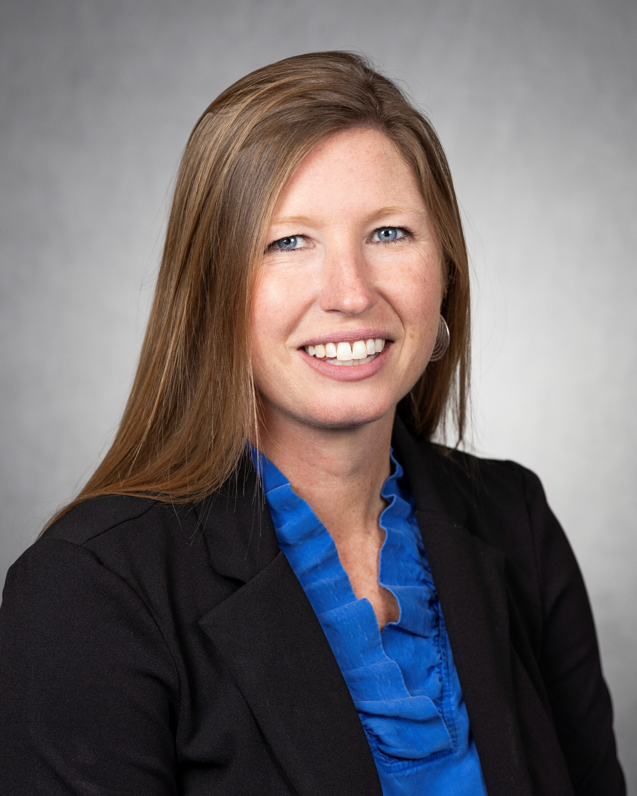 A professional studio headshot of a woman with long, straight light brown hair smiling at the camera. She has blue eyes, light freckles, and is wearing a black blazer over a vibrant blue ruffled top against a solid light gray background.