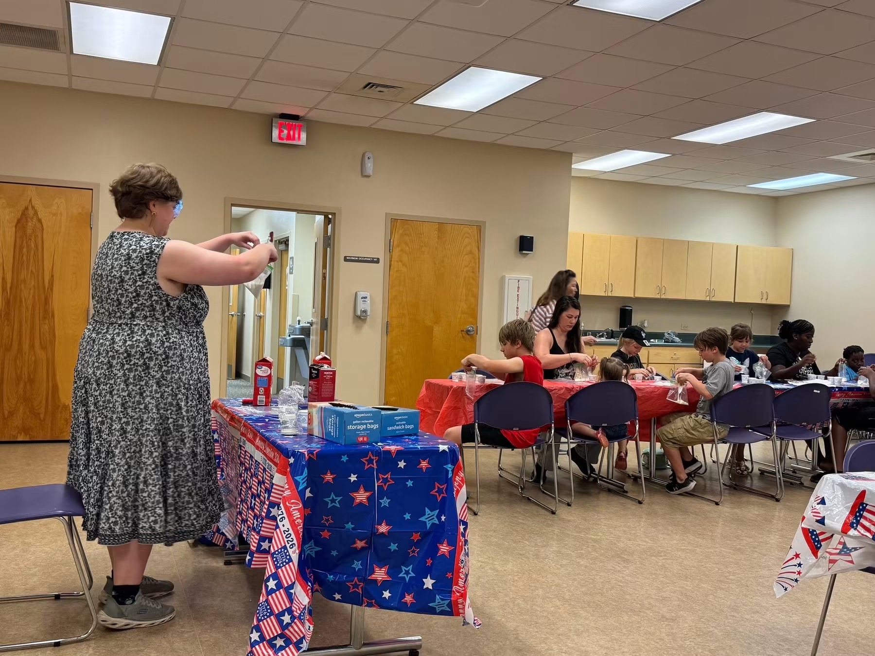 An indoor community room scene where several children and adults are seated at long tables making homemade ice cream. The tables are decorated with red, white, and blue patriotic tablecloths. On the left, an instructor in a patterned dress prepares supplies on a utility table holding milk cartons and a blue box of storage bags.