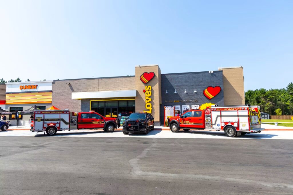 A wide exterior shot of a modern Love's Travel Stop and attached Dunkin' storefront on a clear day. Parked in a line across the asphalt parking lot out front are three emergency vehicles: a red Midway Fire Rescue truck, a black police utility vehicle, and a red City of Gretna Fire Rescue truck.
