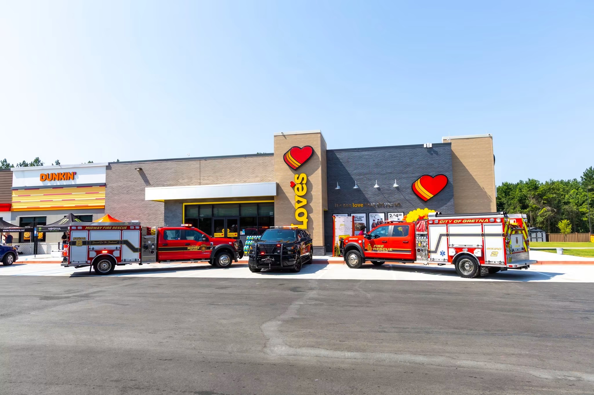 A wide exterior shot of a modern Love's Travel Stop and attached Dunkin' storefront on a clear day. Parked in a line across the asphalt parking lot out front are three emergency vehicles: a red Midway Fire Rescue truck, a black police utility vehicle, and a red City of Gretna Fire Rescue truck.