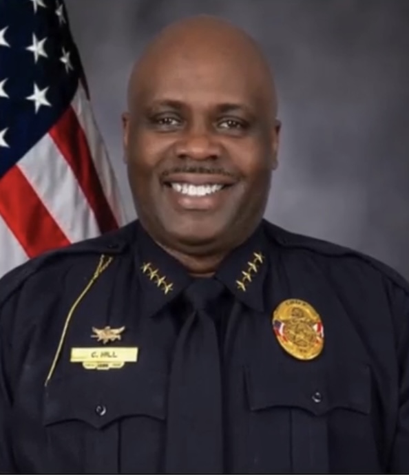 A professional studio headshot of a Black male police officer, Chief C. Hill, smiling broadly at the camera. He is dressed in a dark blue law enforcement uniform featuring four gold stars on each collar flap, a gold badge on his chest, and a gold name tag that reads 'C. HILL' against a gray background with an American flag on the left.